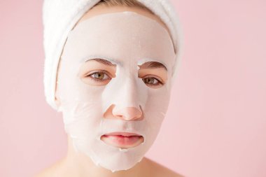 Güzel genç kadın kozmetik doku maske pembe bir arka plan üzerinde bir yüzünde uyguluyor. Sağlık ve güzellik tedavi ve teknoloji kavramı