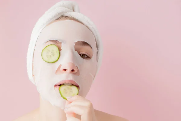 Mulher bonita está aplicando uma máscara de tecido cosmético em um rosto com pepino em um fundo rosa — Fotografia de Stock