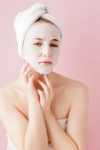 Piękna młoda kobieta jest zastosowanie kosmetycznych tkanek maski na twarz na różowym tle. Opieki zdrowotnej i piękna koncepcja leczenia i technologii — Zdjęcie stockowe