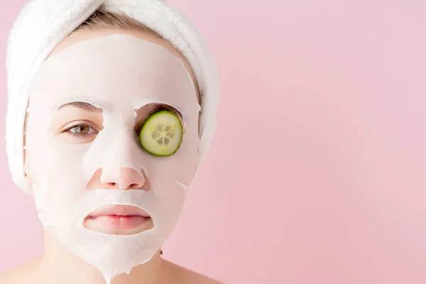 Bela jovem está aplicando uma máscara de tecido cosmético em um rosto com pepino em um fundo rosa. Cuidados de saúde e beleza e conceito de tecnologia — Fotografia de Stock