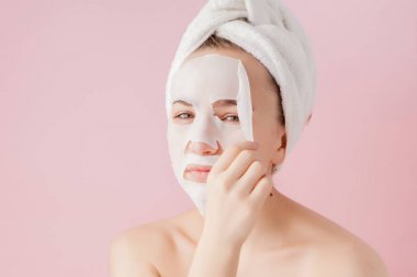 Güzel genç kadın kozmetik doku maske pembe bir arka plan üzerinde bir yüzünde uyguluyor. Sağlık ve güzellik tedavi ve teknoloji kavramı