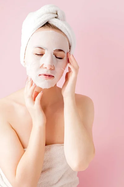 Bela jovem está aplicando uma máscara de tecido cosmético em um rosto em um fundo rosa. Cuidados de saúde e beleza e conceito de tecnologia — Fotografia de Stock
