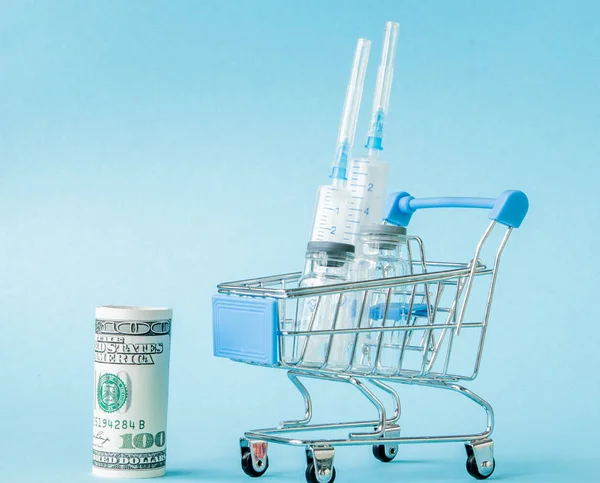 Medical injection and dollars in shopping trolley on blue background. Creative idea for health care cost, drugstore, health insurance and pharmaceutical company business concept. Copy space