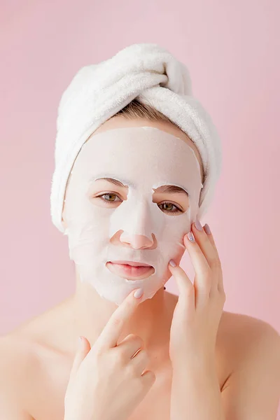 Hermosa mujer joven está aplicando una máscara de tejido cosmético en una cara sobre un fondo rosa. Salud y belleza tratamiento y concepto de tecnología —  Fotos de Stock