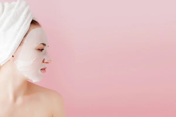 Wanita muda yang cantik menerapkan masker jaringan kosmetik pada wajah dengan latar belakang merah muda. Kesehatan dan kecantikan pengobatan dan konsep teknologi — Stok Foto