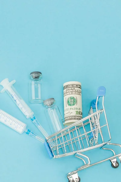 Medical injection and dollars in shopping trolley on blue backgr