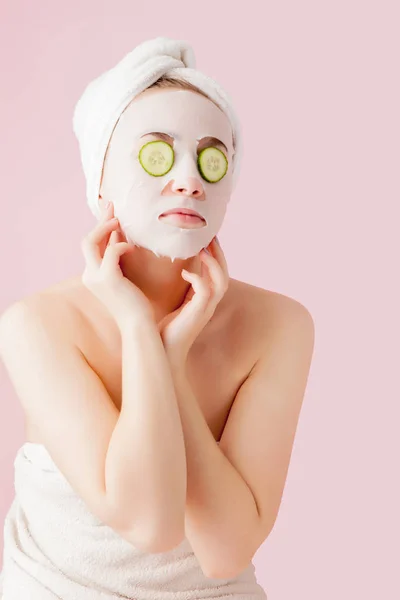 Krásná mladá žena je použití kosmetické tkáňové masky na tváři s okurkou na růžovém pozadí — Stock fotografie