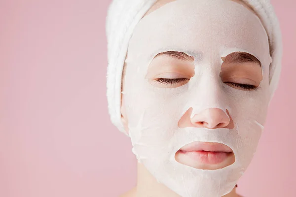 Belle jeune femme applique un masque de tissu cosmétique sur un visage sur un fond rose. Soins de santé et soins de beauté et concept technologique — Photo