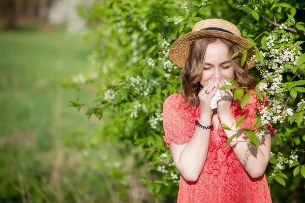 花の木の前で鼻を吹いてティッシュでくしゃみをする若い女の子。季節性アレルゲンが人々に影響を与えます。美しい女性は鼻炎を持っている. — ストック写真
