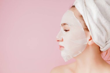Güzel genç kadın kozmetik doku maske pembe bir arka plan üzerinde bir yüzünde uyguluyor. Sağlık ve güzellik tedavi ve teknoloji kavramı