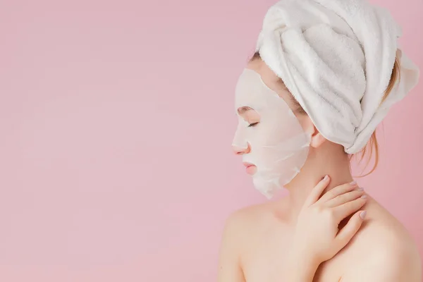 Hermosa mujer joven está aplicando una máscara de tejido cosmético en una cara sobre un fondo rosa. Salud y belleza tratamiento y concepto de tecnología — Foto de Stock