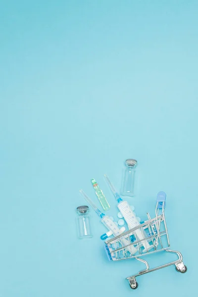 Pills and medical injection in shopping trolley on blue backgrou