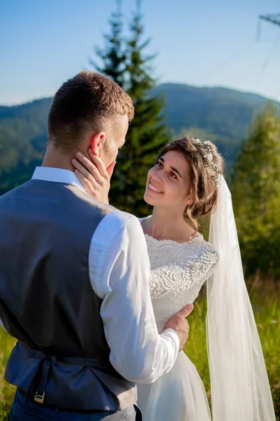 Frischvermählte lächeln und umarmen sich auf der Wiese auf dem Gipfel des Berges. Hochzeitsspaziergang im Wald in den Bergen, die sanften Emotionen des Paares, Foto zum Valentinstag — Stockfoto