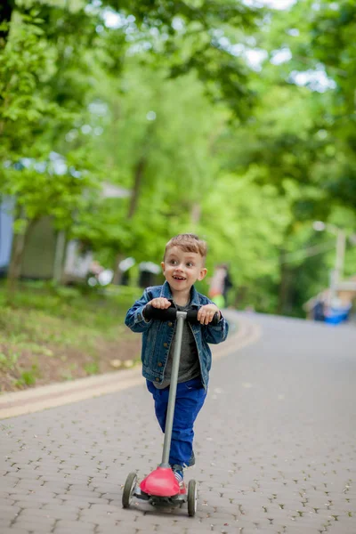 Menino montando scooter no parque da cidade em aummer. Crianças desportos ao ar livre. Criança feliz brincando com sua scooter. O miúdo aprende a andar de scooter no parque. Infância feliz — Fotografia de Stock
