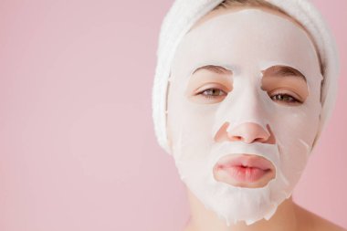 Güzel genç kadın kozmetik doku maske pembe bir arka plan üzerinde bir yüzünde uyguluyor