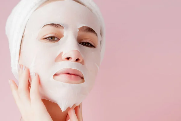 Wanita muda yang cantik menerapkan masker jaringan kosmetik pada wajah pada latar belakang merah muda — Stok Foto