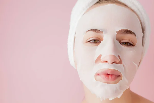 Hermosa mujer joven está aplicando una máscara de tejido cosmético en una cara sobre un fondo rosa — Foto de Stock