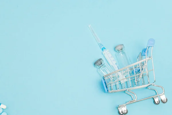 Medical injection in shopping trolley on blue background. Creative idea for health care cost, drugstore, health insurance and pharmaceutical company business concept. Copy space