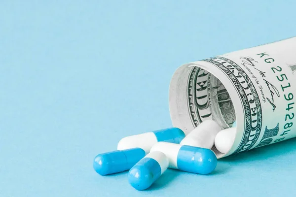 Dinheiro dólar enrolado com pílulas fluindo isolado em fundo azul, altos custos de conceito de medicação caro. Espaço de cópia — Fotografia de Stock