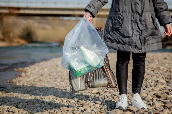 Σκουπίδια στη φύση, καθαρισμός του περιβάλλοντος την άνοιξη στον ποταμό από τα σκουπίδια μια γυναίκα σε μπλε γάντια λατέξ μίας χρήσης σε μπλε μεγάλη πλαστική σακούλα — Φωτογραφία Αρχείου