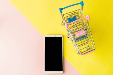 Pastel pembe ve sarı arka planda boş ekran beyaz cep telefonu ve alışveriş arabaları. Minimal stil, flatlay