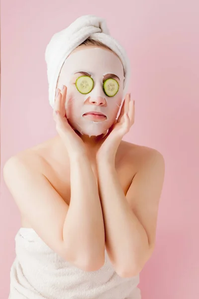 Belle jeune femme applique un masque de tissu cosmétique sur un visage avec concombre sur un fond rose — Photo