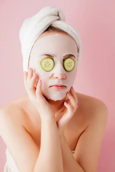 Wanita muda yang cantik menerapkan masker jaringan kosmetik pada wajah dengan mentimun pada latar belakang merah muda — Stok Foto