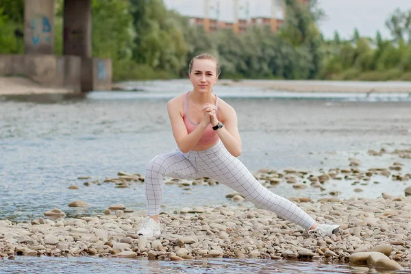 Girl in sports uniforms makes a stretch on the river bank — Stock Photo, Image