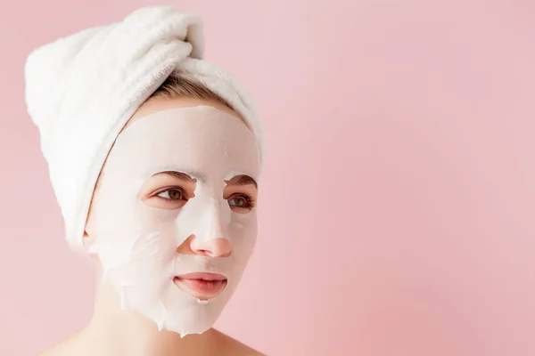 Hermosa mujer joven está aplicando una máscara de tejido cosmético en una cara sobre un fondo rosa — Foto de Stock