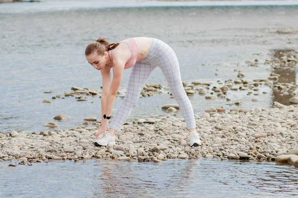 Girl in sports uniforms makes a stretch on the river bank