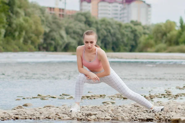 Girl in sports uniforms makes a stretch on the river bank