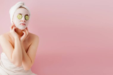 Güzel genç bir kadın pembe arka planda salatalıklı bir yüze kozmetik doku maskesi takıyor.