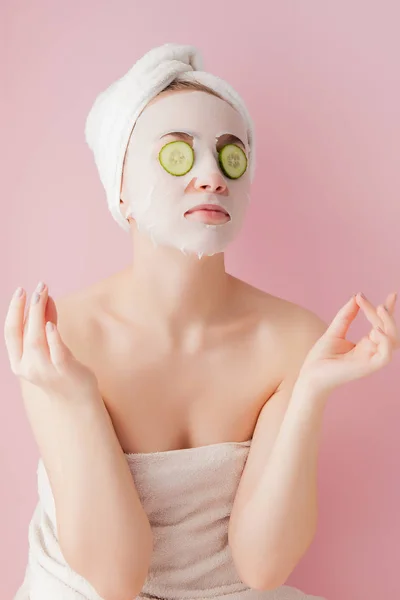 Belle jeune femme applique un masque de tissu cosmétique sur un visage avec concombre sur un fond rose — Photo