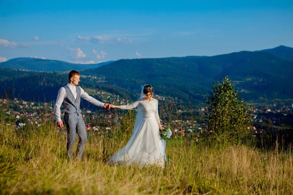 Novomanželé úsměv a objetí vzájemně mezi na louce na vrcholu hory. Svatební procházka v lese v horách, jemné emoce páru, Foto pro den svatého Valentýna — Stock fotografie