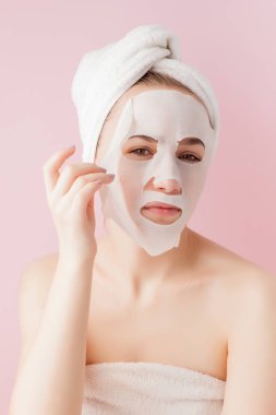 Güzel genç kadın kozmetik doku maske pembe bir arka plan üzerinde bir yüzünde uyguluyor