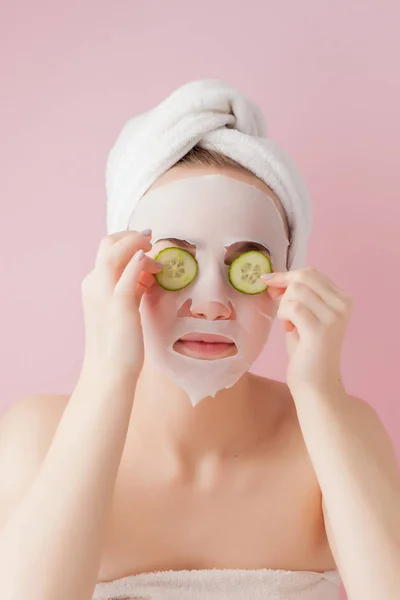 Hermosa mujer joven está aplicando una máscara de tejido cosmético en una cara con pepino sobre un fondo rosa. Salud y belleza tratamiento y concepto de tecnología — Foto de Stock