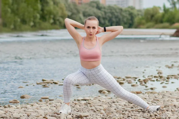 Girl in sports uniforms makes a stretch on the river bank