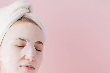 Güzel genç kadın kozmetik doku maske pembe bir arka plan üzerinde bir yüzünde uyguluyor. Sağlık ve güzellik tedavi ve teknoloji kavramı
