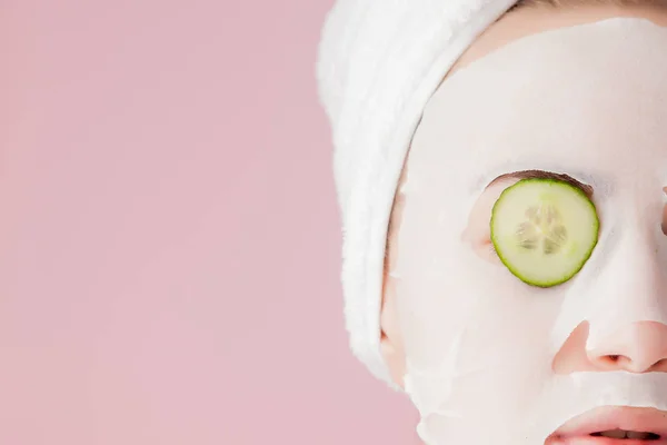 Mooie jonge vrouw is het aanbrengen van een cosmetische weefsel masker op een gezicht met komkommer op een roze achtergrond — Stockfoto