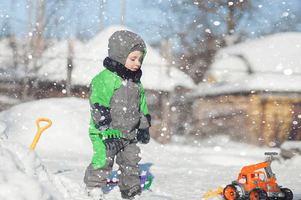 Porträt eines niedlichen Kleinkindes, das auf Schnee sitzt und mit seinem gelben Traktorspielzeug im Park spielt. Kind spielt im Freien. Glücklicher Junge mit Bauspielzeug. Lifestylekonzept — Stockfoto