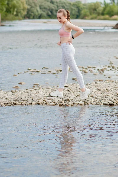 Girl in sports uniforms makes a stretch on the river bank