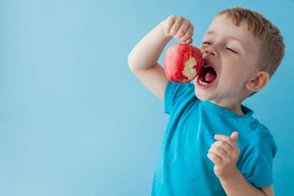 Ребенок держит и ест красное яблоко на голубом фоне, питание, диета и здоровое питание — стоковое фото