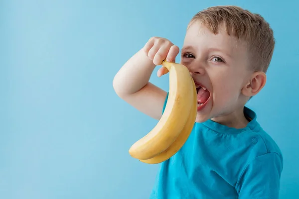 Little Boy Sostener y comer un plátano sobre fondo azul, comida, dieta y concepto de alimentación saludable — Foto de Stock