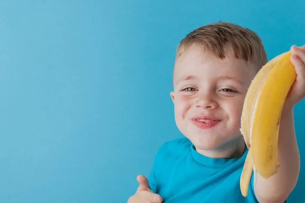 Little Boy Segurando e comendo uma banana em fundo azul, comida, dieta e conceito de alimentação saudável — Fotografia de Stock