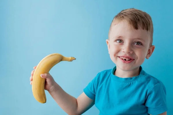 Little Boy Segurando e comendo uma banana em fundo azul, comida, dieta e conceito de alimentação saudável — Fotografia de Stock