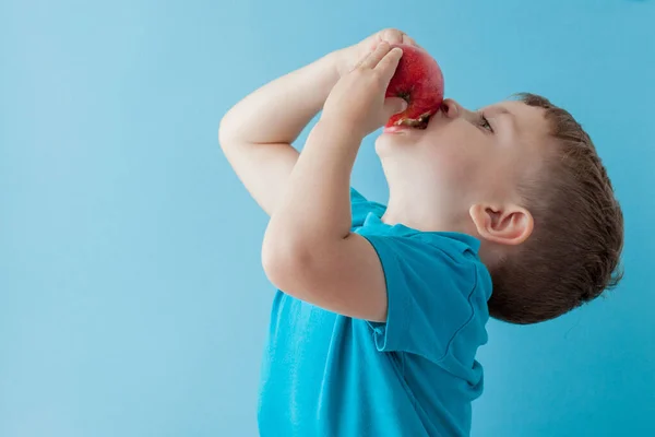 Bebé niño sosteniendo y comiendo manzana roja sobre fondo azul, comida — Foto de Stock