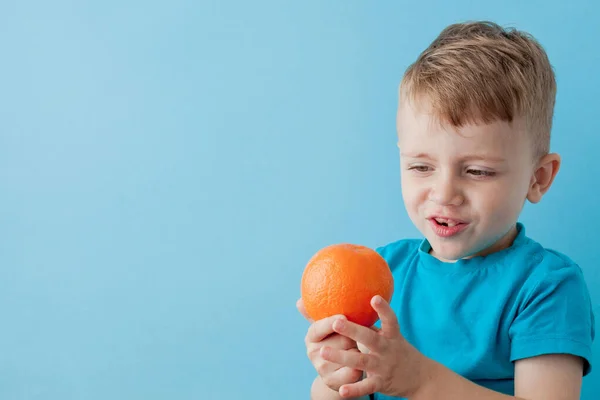 Niño pequeño sosteniendo una naranja en sus manos sobre fondo azul, di — Foto de Stock