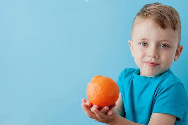 Menino segurando uma laranja em suas mãos no fundo azul, di — Fotografia de Stock