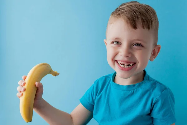 Little Boy Segurando e comendo uma banana em fundo azul, comida, dieta e conceito de alimentação saudável — Fotografia de Stock