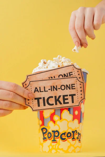 Hand tar en biobiljett och popcorn från en papperskopp på en gul bakgrund. Kvinnan äter popcorn. Filmkoncept. Ett platt ligg. Kopiera utrymme — Stockfoto